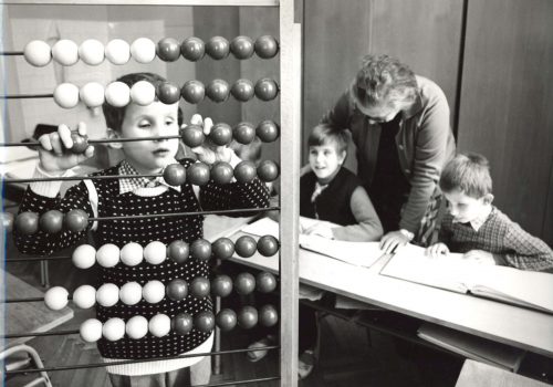 Nevidiaci žiak počítajúci na počítadle. V pozadí ďalší žiaci s pani učiteľkou Repaskou. Rok 1986.