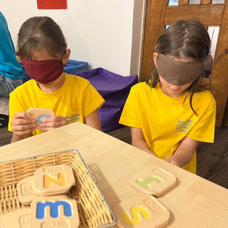 Malí múzejníci s maskami na očiach hmatajú písmená.